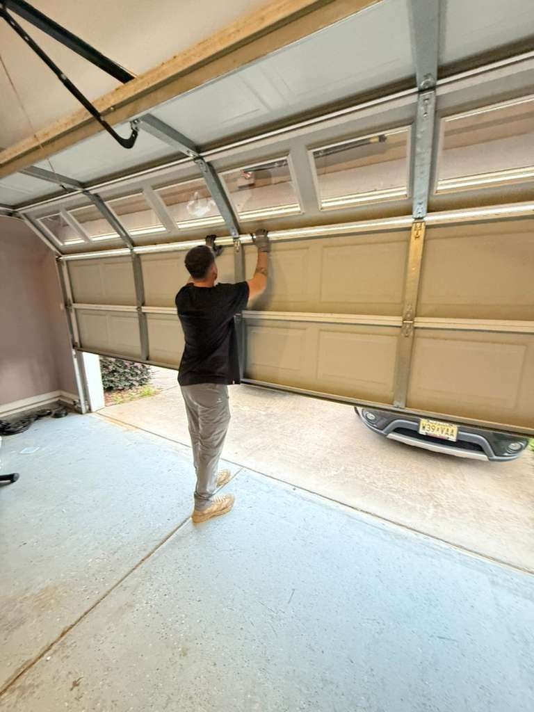 Technician in a black shirt holding a sectional garage door halfway up by hand, with a car parked in the driveway outside
