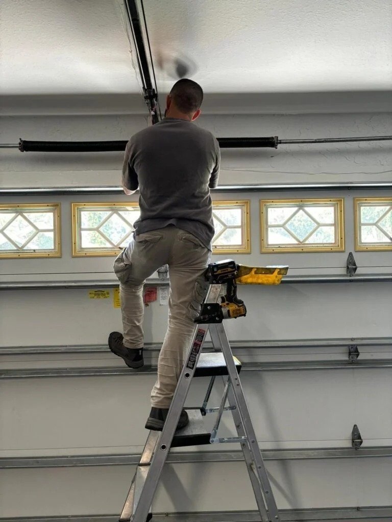 Technician on a stepladder with a cordless drill working at the top of a garage door that has diamond-pattern window panels