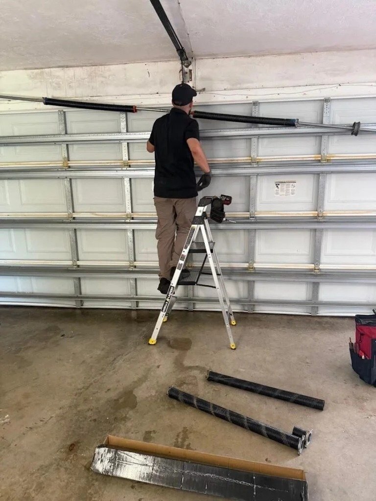 Technician on a stepladder at the torsion spring above a white garage door, with new steel parts laid out on the floor