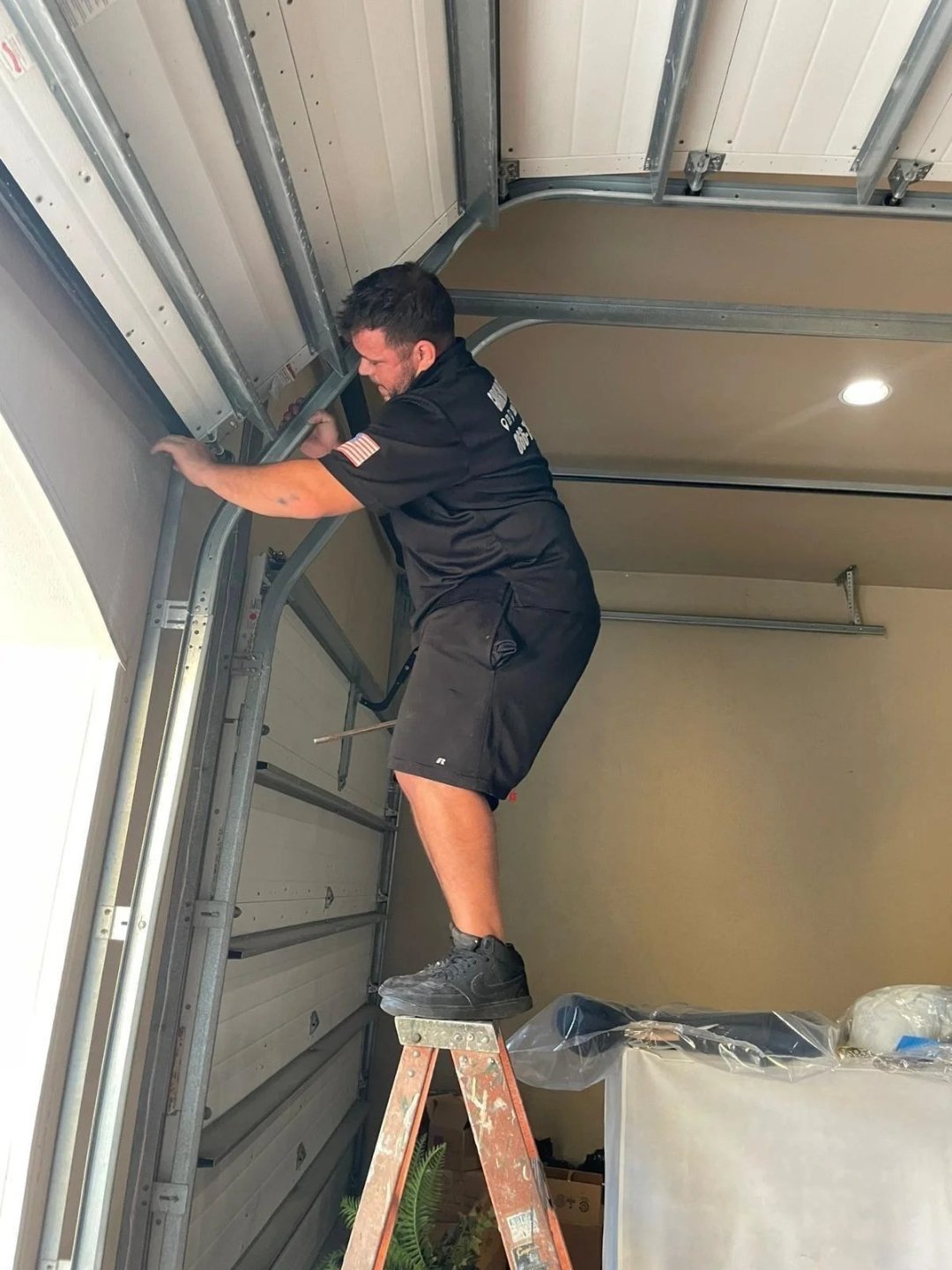 Technician in black shorts on a ladder holding the vertical track and bracket at the side of a garage door