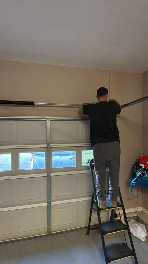 A technician on a stepladder at the ceiling of a garage with a door that has windowed sections